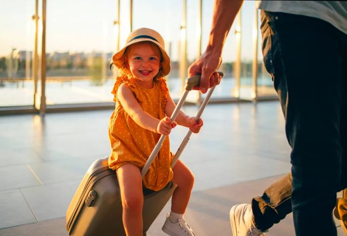 A little girl joyfully riding on top of a suitcase while a man pulls it across a tiled floor.