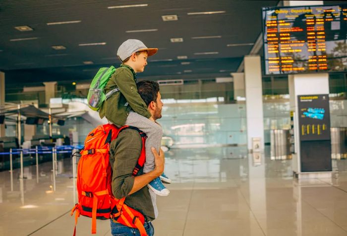 A man with an orange bag is carrying a child on his shoulders while standing in an airport.