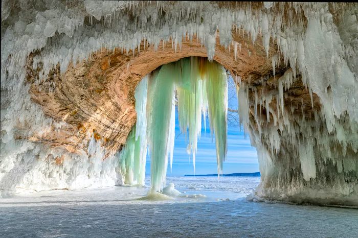 Brilliant white stalactites framing a cave entrance, with a clear blue sky and snowy landscape visible through the opening