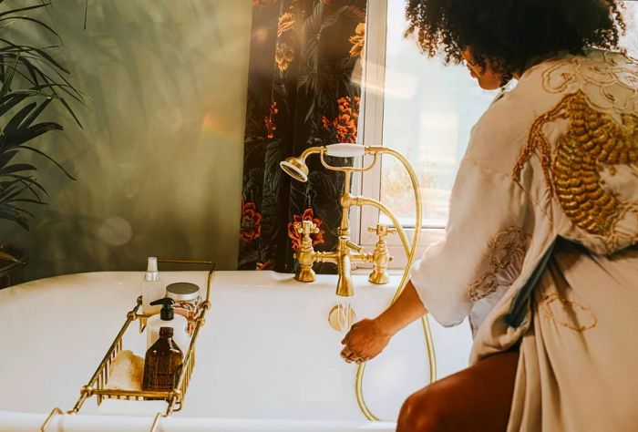 A woman with curly hair, wrapped in a bathrobe, tests the temperature of the water in the bathtub.