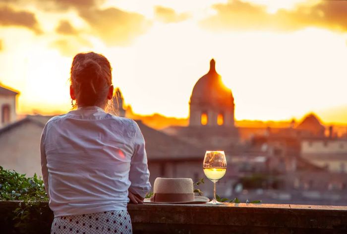 A woman rests against a balcony railing beside a hat and a glass of wine, watching the sunset over the city.