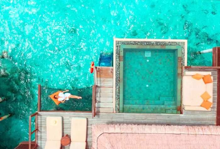 An overwater villa featuring a woman lounging on a net bed next to a square pool equipped with a couch and sun loungers.