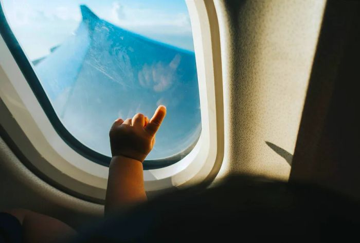 A toddler excitedly points at an airplane window, gazing at a cloudy blue sky.