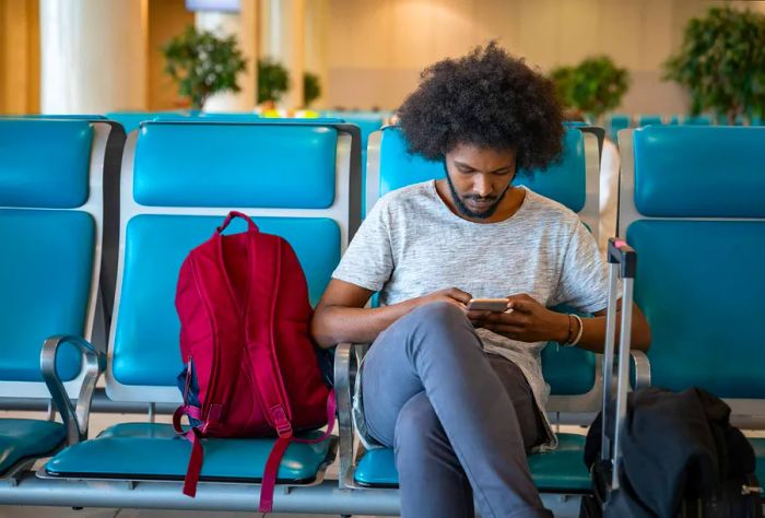 A male traveler is seen at the airport, smiling while using his cell phone as he waits for his flight.