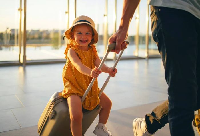 An anonymous father walks with his young daughter sitting on a piece of luggage, smiling at the camera in the airport.