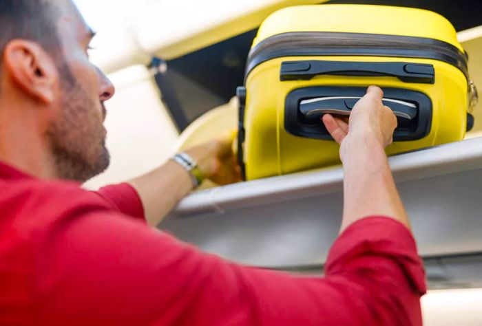 A traveler retrieving their hand luggage from the overhead compartment on a plane.