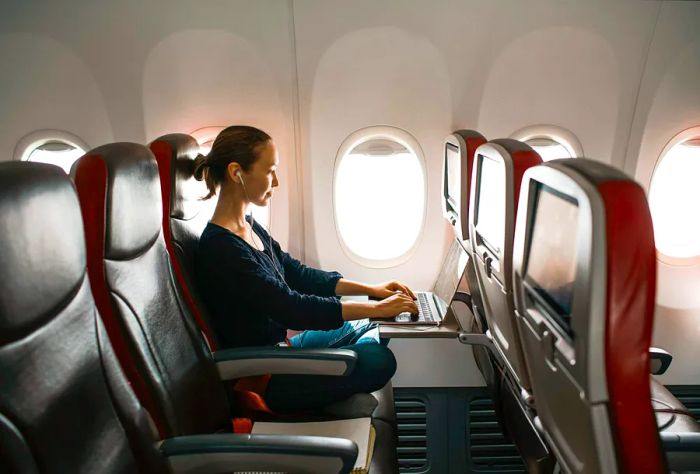 A woman seated by the window in an aircraft, working on her laptop.