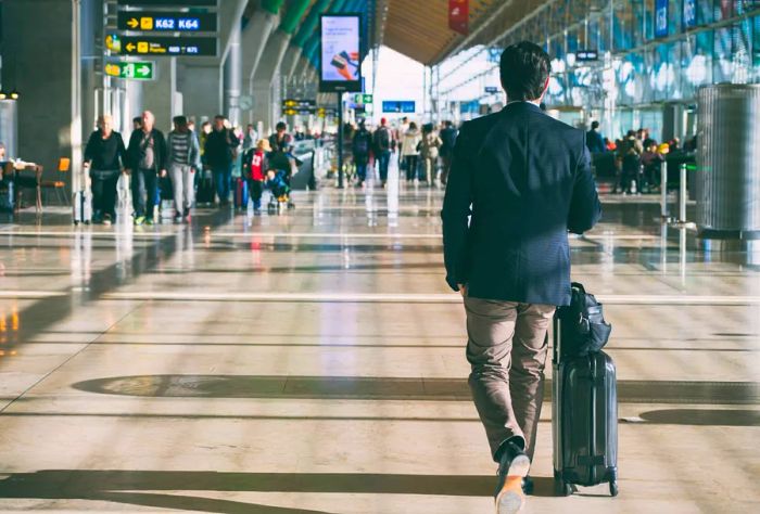 A view from behind of a man with a suitcase and various other travelers walking through the airport corridor.