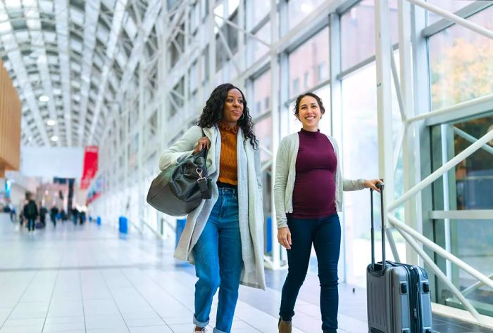 Two female friends are traveling together. One woman is black, while the other is Eurasian and pregnant. Both smiling women are happily chatting as they carry their luggage, walking side by side through the airport—an embodiment of the babymoon and travel safety during pregnancy.