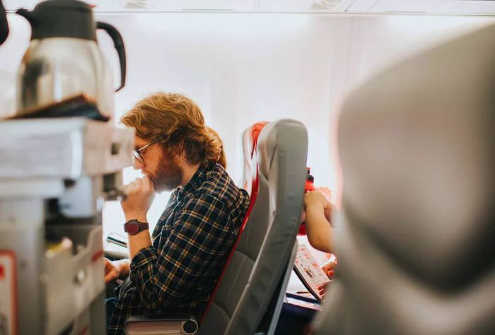 A man sits comfortably on a plane, dressed in checkered long sleeves and glasses, deeply engrossed in a book.