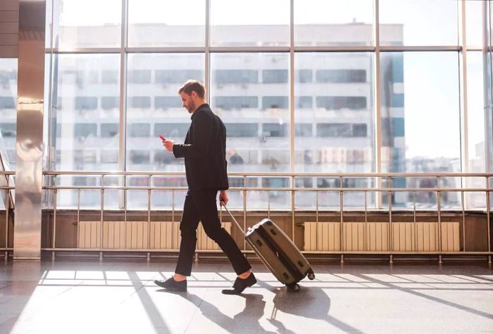 A male traveler checks his smartphone while navigating the airport with his luggage.