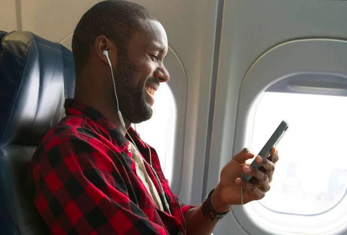 A smiling male passenger wearing earphones, gazing at his smartphone from the window seat of the plane.