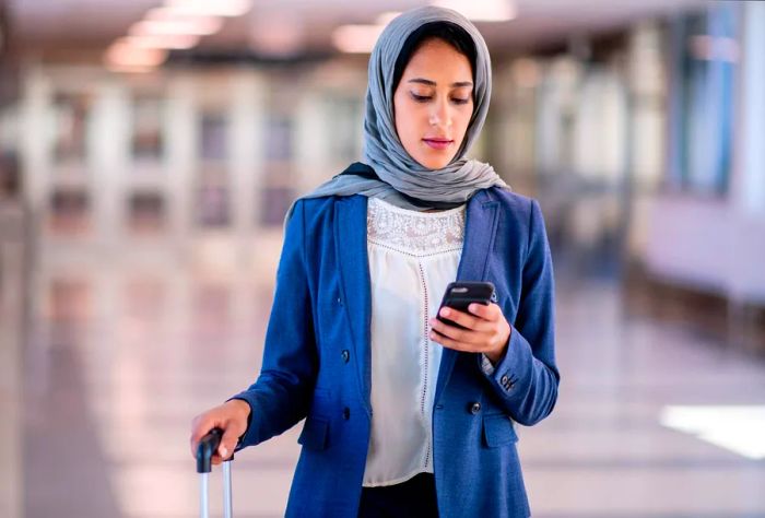 A woman wearing a hijab checks her mobile phone while holding a suitcase.