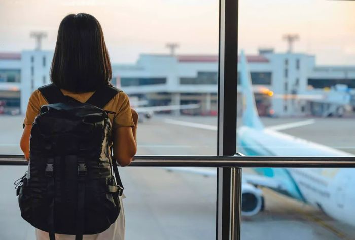 A traveler stands with luggage, watching the sunset from an airport window. A woman gazes at the lounge while observing planes, waiting at the boarding gate for her departure.