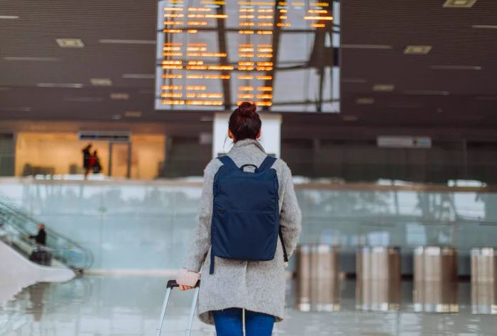 A woman at the airport, looking at the flight information boards while holding her suitcase and wearing a backpack.