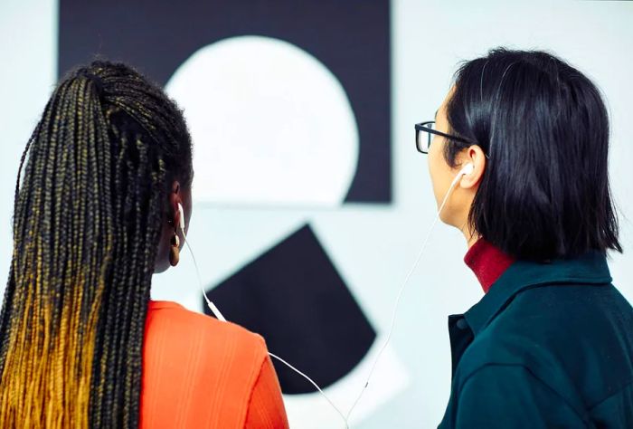 A man with dark hair and a woman sporting cornrow braids share a headphone as they admire a piece of wall art.