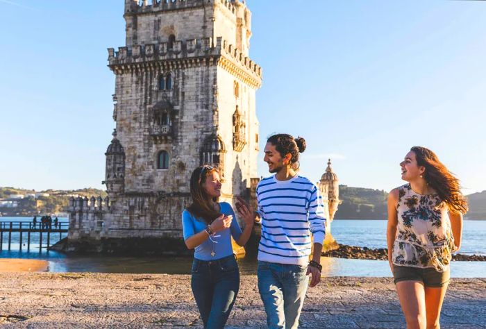 A trio of young people strolls along a riverside promenade, with Belém Tower in the background.