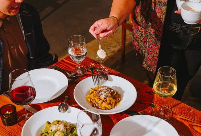 A woman sprinkles parmesan cheese over a plate of pasta at a customer's table.