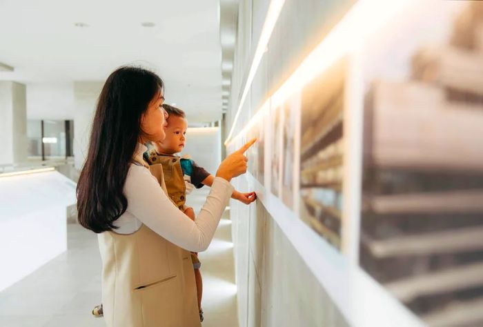 A woman points to a painting on the wall while holding a small child.