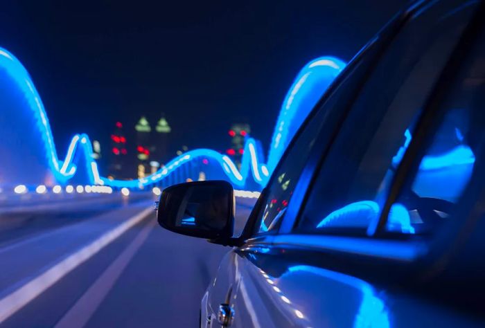 A car travels over a neon-lit bridge at night, with lights shimmering on its surface.