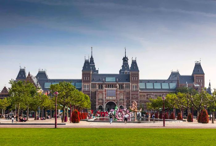 Gothic architecture features a grand museum front, framed by tall green trees and a central pond adorned with a large rope-like sculpture.