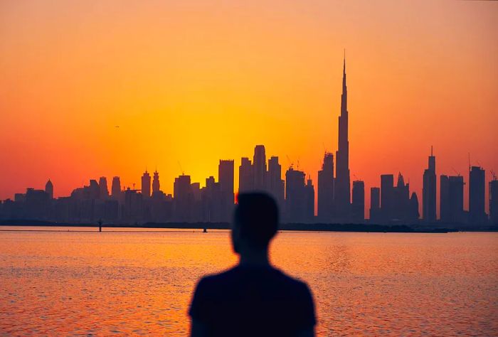 A silhouette of an individual gazes at the urban skyline by the sea, with modern skyscrapers set against a stunning twilight sky.