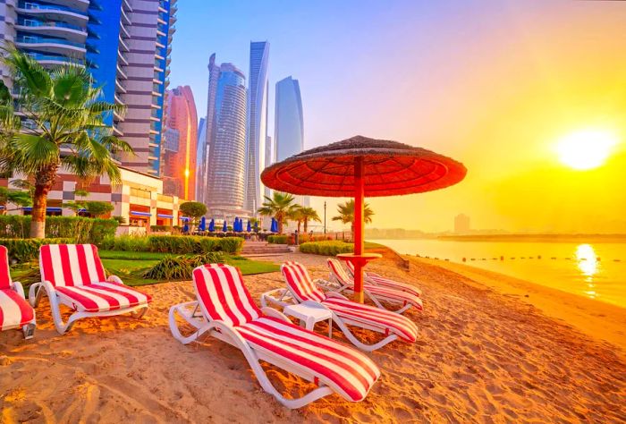 At dawn, a row of red-and-white striped sunbeds sits beneath a hut umbrella on a pristine sandy beach, framed by towering buildings.