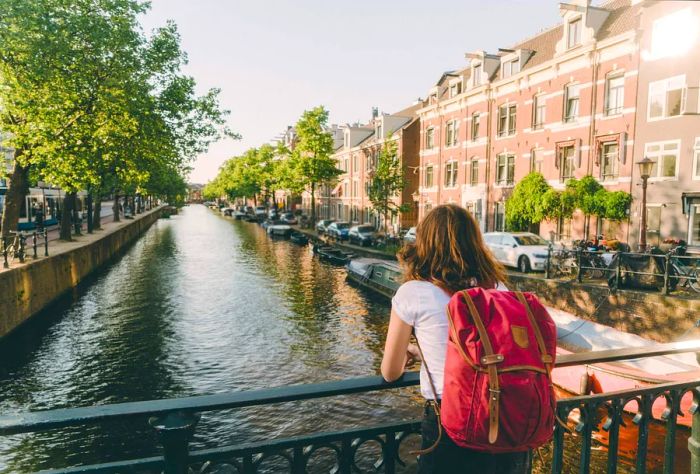 A woman with a backpack leans against the railing of a bridge, gazing thoughtfully over a canal.