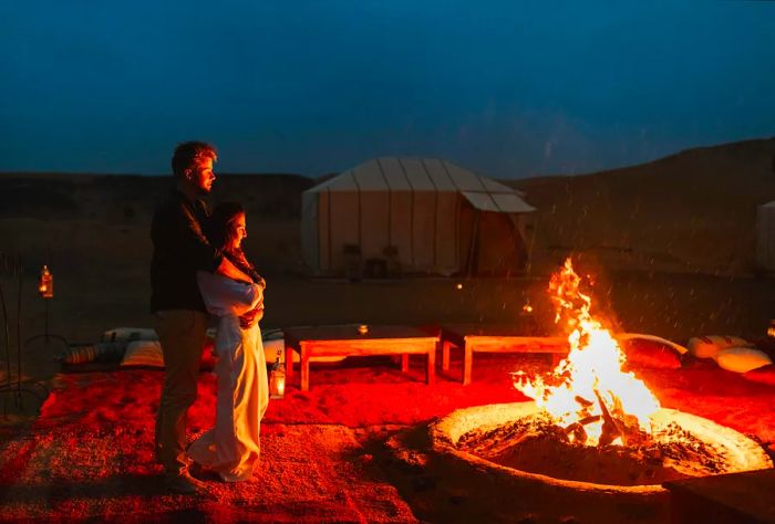 A couple enjoys a cozy campfire in the desert, with one person embracing the other from behind.