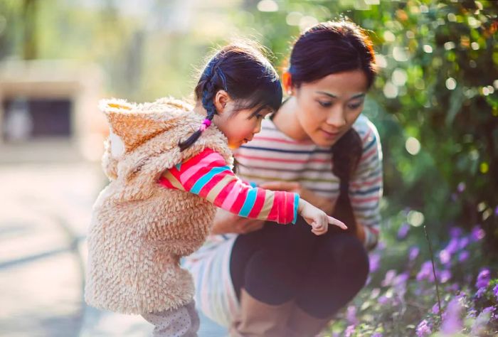 A lovely toddler girl excitedly points out flowers to her beautiful young mom along a park walkway.