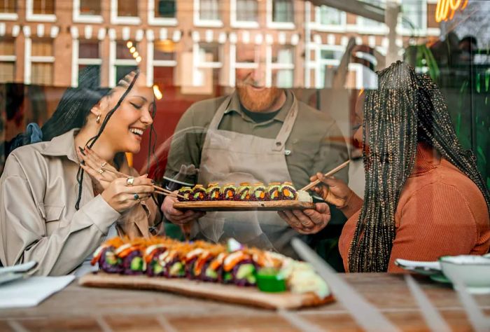 A senior male staff member serves sushi on wooden boards to two female diners inside the restaurant.