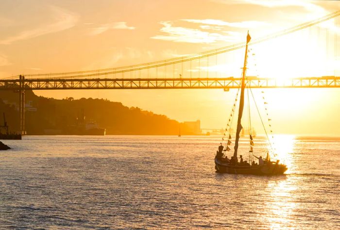 A ship gliding across a river beneath a suspension bridge at sunset.