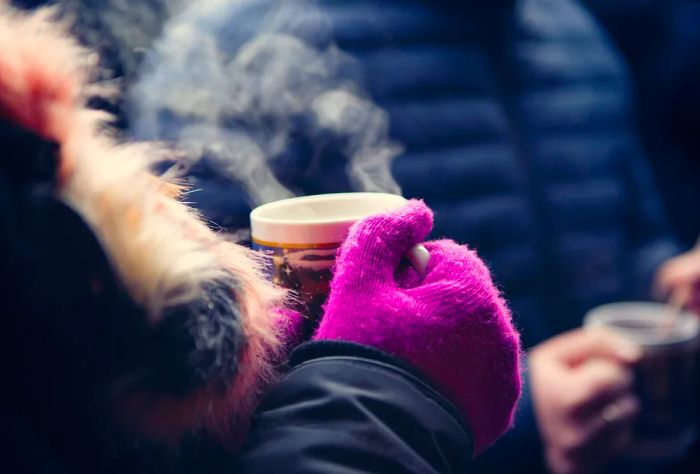 A person in a winter jacket and purple gloves enjoys a steaming mug of hot beverage.