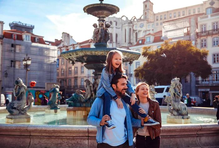 A man hoists a child onto his shoulders while standing next to a woman, with a grand fountain in the background.