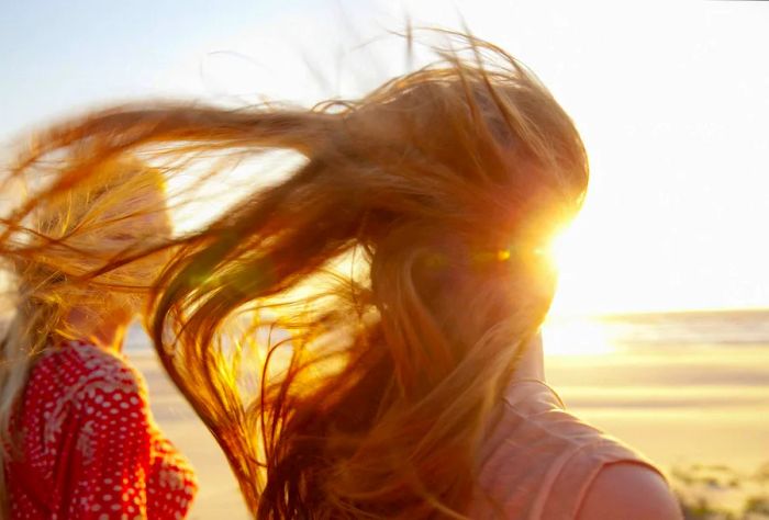 A woman with flowing golden hair stands beside another woman on the beach, her hair dancing in the breeze.