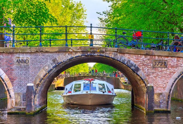 A canal boat carrying passengers is about to pass under an arched bridge.