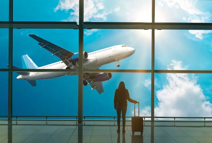 A young woman stands in the airport's departure hall, watching an airplane take off outside while holding a suitcase.