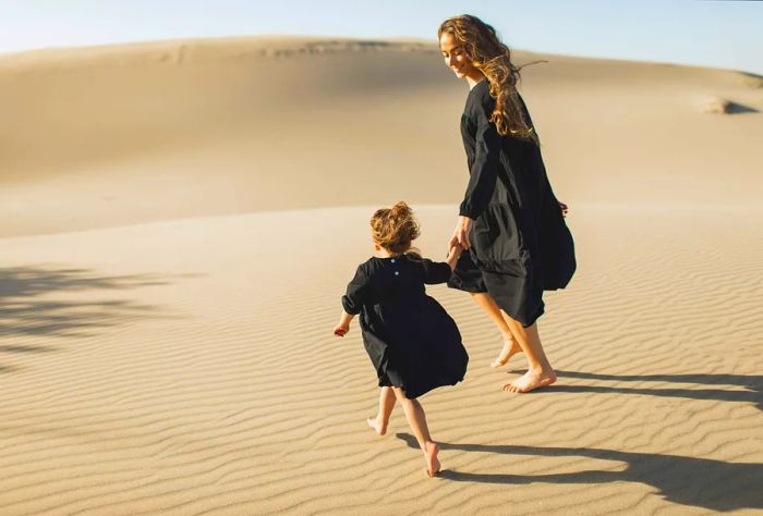 A mother and daughter wander barefoot across the sand dunes in the desert.