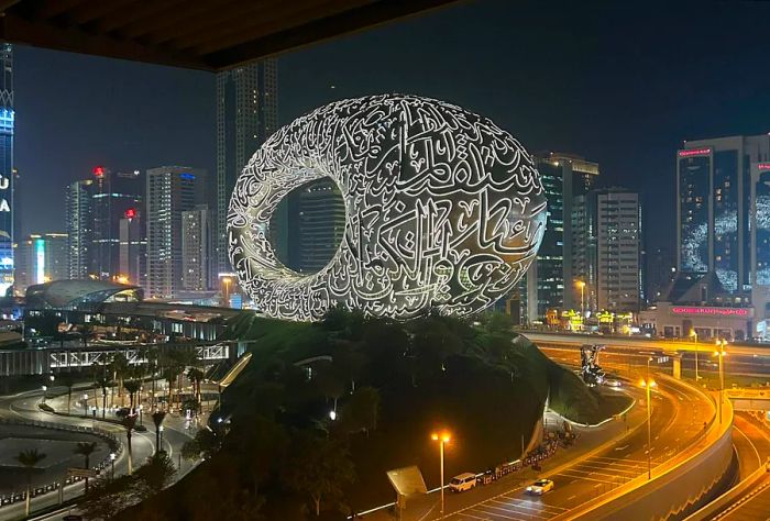 A stunning monument shaped like a sphere, adorned with bright LED Arabic letters, stands alongside the road.