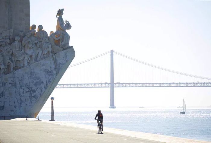 A cyclist approaches a riverside monument featuring statues of figures ascending a slope, framed by a suspension bridge in the background.