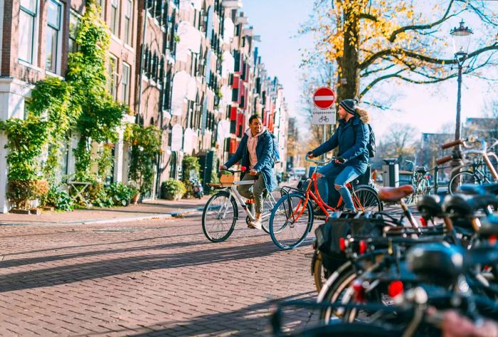 A joyful couple enjoys their city getaway as they cycle along a sidewalk, passing a picturesque row of buildings and parked bicycles.