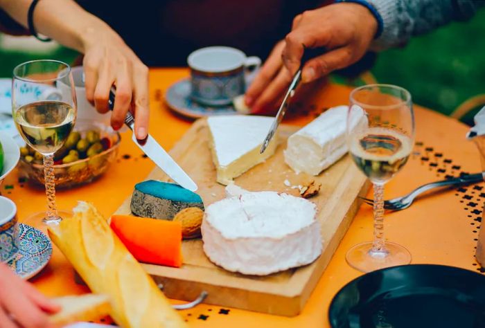 Guests gather around tables filled with an assortment of cheeses, wines, baguette, fruits, and vegetables.