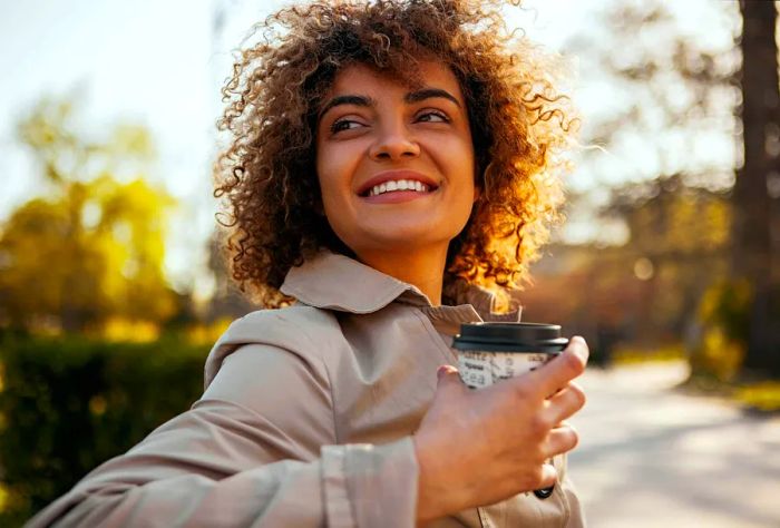 Close-up of a cheerful woman savoring her takeaway coffee on a bench in a park during autumn.