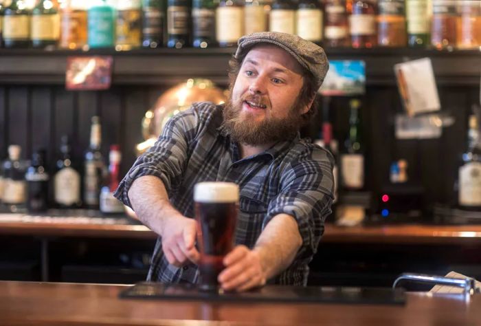 A bartender, wearing a knowing smile, points to a freshly made drink on the counter while scanning the crowd for the next customer.