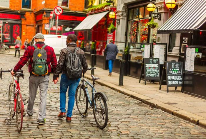 A gay couple dressed for winter, holding hands and pulling their bikes along the cobblestone street in front of shops.