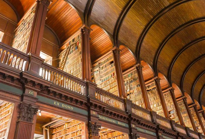 A library's upper arcade lined with wooden shelves brimming with books and equipped with rolling ladders.