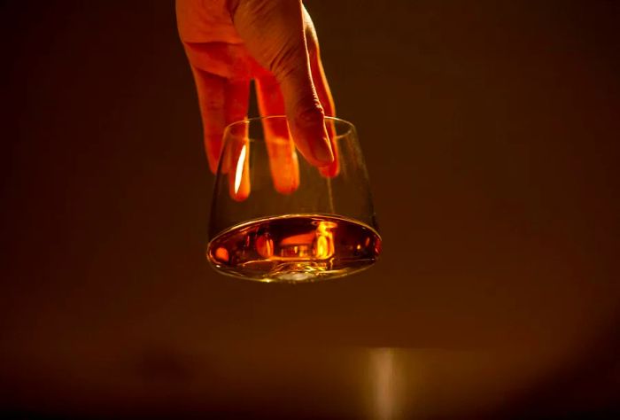 A hand holds a clear glass filled with golden liquor against a warm brown background.