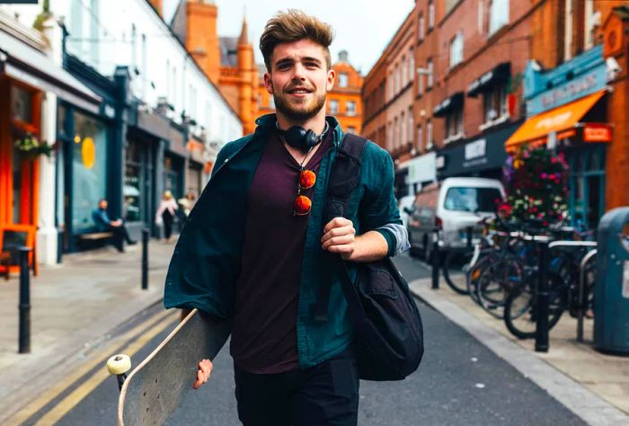 A fashionable young man strolls down a street, holding a bag and a skateboard.