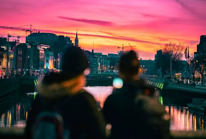 Silhouetted against the city skyline, two indistinct figures can be spotted standing on a bridge above the river.