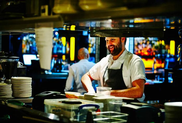 A smiling chef works behind a counter filled with neatly stacked dishes in the kitchen.